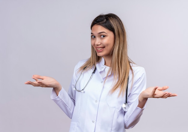 Hermosa joven médico vistiendo bata blanca con estetoscopio mirando positivo y feliz sonriendo extendiendo las palmas hacia los lados