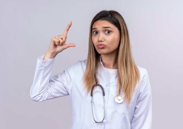 Hermosa joven médico vistiendo bata blanca con estetoscopio mirando preocupado haciendo signo de tamaño con los dedos símbolo de medida