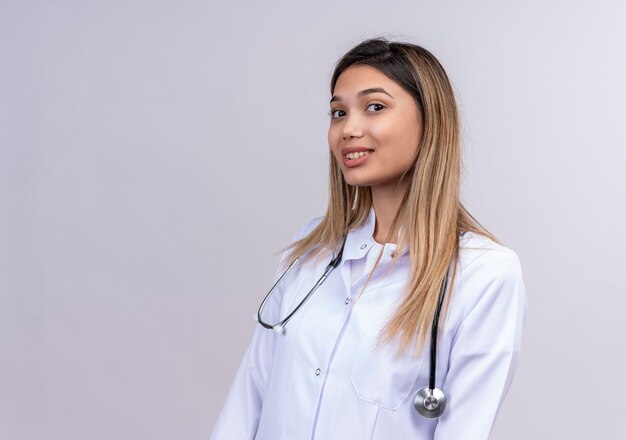 Hermosa joven médico vistiendo bata blanca con estetoscopio mirando con sonrisa de confianza en la cara