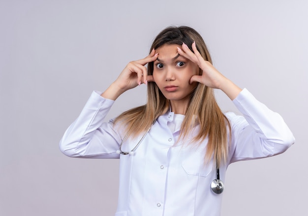 Hermosa joven médico vistiendo bata blanca con estetoscopio mirando tratando de abrir los ojos con los dedos adormilados y cansados por la fatiga matutina