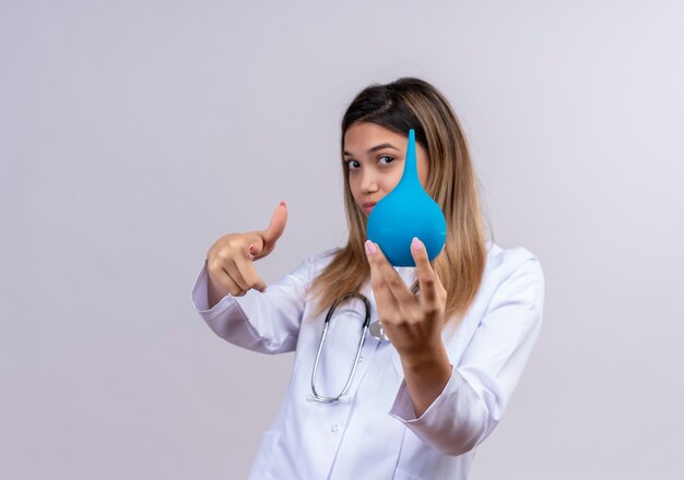 Hermosa joven médico vistiendo bata blanca con estetoscopio mostrando un enema apuntando con el dedo índice al frente