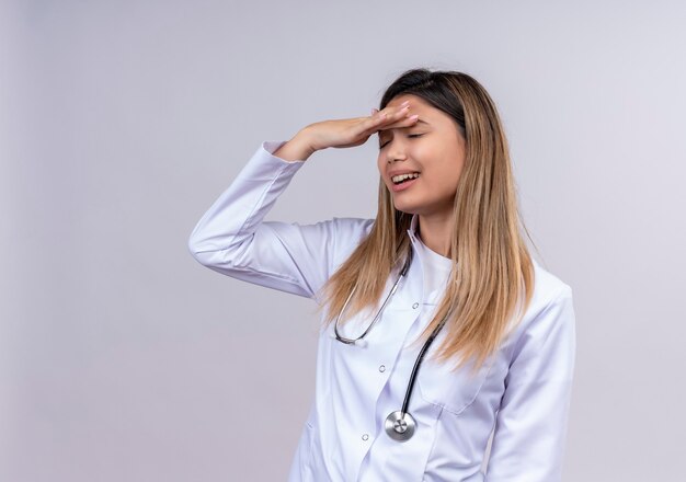 Hermosa joven médico vistiendo bata blanca con estetoscopio de pie con la mano en la cabeza por error mirando confundido