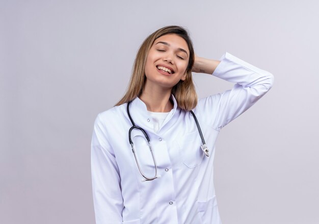 Hermosa joven médico vistiendo bata blanca con estetoscopio de pie con los ojos cerrados estirando ella misma