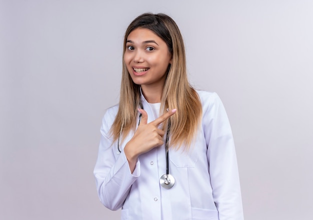 Hermosa joven médico vistiendo bata blanca con estetoscopio sonriendo alegremente apuntando con el dedo índice hacia el lado