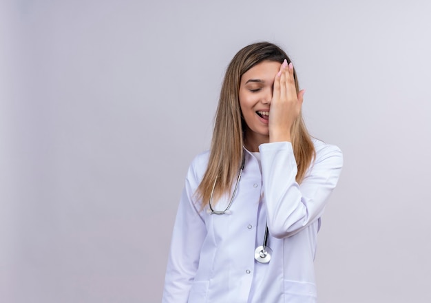 Hermosa joven médico vistiendo bata blanca con estetoscopio sonriendo alegremente cubriendo los ojos con la mano