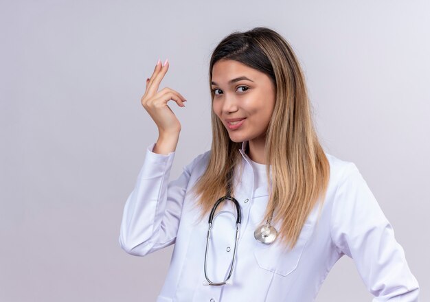 Hermosa joven médico vistiendo bata blanca con estetoscopio sonriendo alegremente gesticulando con la mano