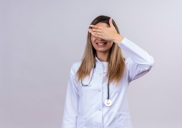 Foto gratuita hermosa joven médico vistiendo bata blanca con estetoscopio sonriendo cubriendo los ojos con el brazo