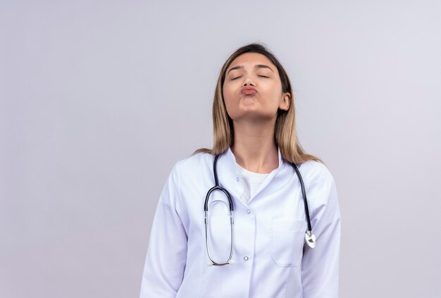 Hermosa joven médico vistiendo bata blanca con estetoscopio soplando mejillas