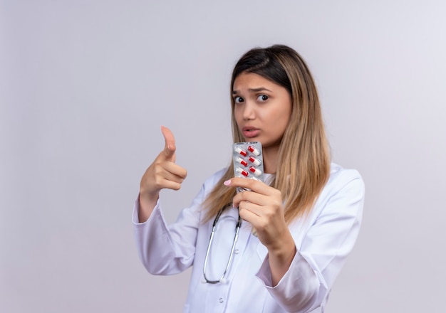 Hermosa joven médico vistiendo bata blanca con estetoscopio sosteniendo blister con pastillas mirando preocupado apuntando con el dedo índice al frente