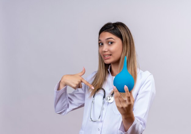 Hermosa joven médico vistiendo bata blanca con estetoscopio sosteniendo un enema apuntando con el dedo hacia él mirando confiado