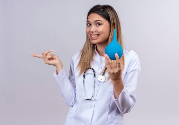 Hermosa joven médico vistiendo bata blanca con estetoscopio sosteniendo un enema sonriendo alegremente apuntando con el dedo hacia el lado
