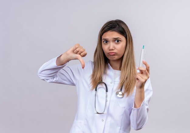 Hermosa joven médico vistiendo bata blanca con estetoscopio sosteniendo una jeringa mirando con el ceño fruncido mostrando los pulgares hacia abajo