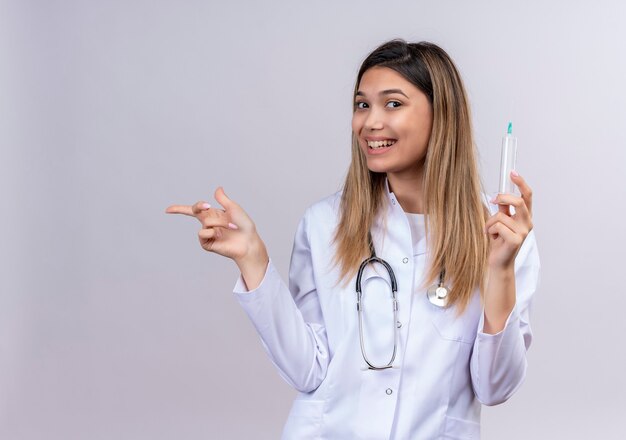 Hermosa joven médico vistiendo bata blanca con estetoscopio sosteniendo una jeringa sonriendo alegremente apuntando con el dedo índice hacia el lado