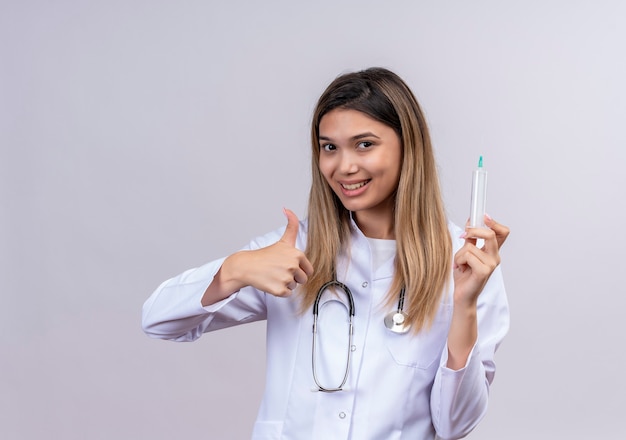 Hermosa joven médico vistiendo bata blanca con estetoscopio sosteniendo jeringa sonriendo alegremente mostrando los pulgares para arriba