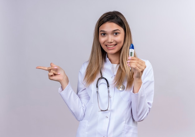 Hermosa joven médico vistiendo bata blanca con estetoscopio sosteniendo termómetro digital sonriendo alegremente apuntando con el dedo índice hacia el lado