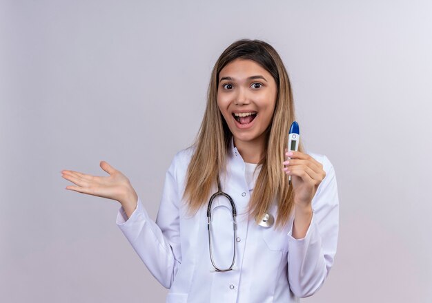 Hermosa joven médico vistiendo bata blanca con estetoscopio sosteniendo termómetro digital sonriendo alegremente presentando con brazo oh su mano