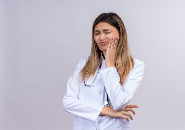 Hermosa joven médico vistiendo bata blanca con estetoscopio tocando su mejilla con aspecto de malestar teniendo dolor de muelas