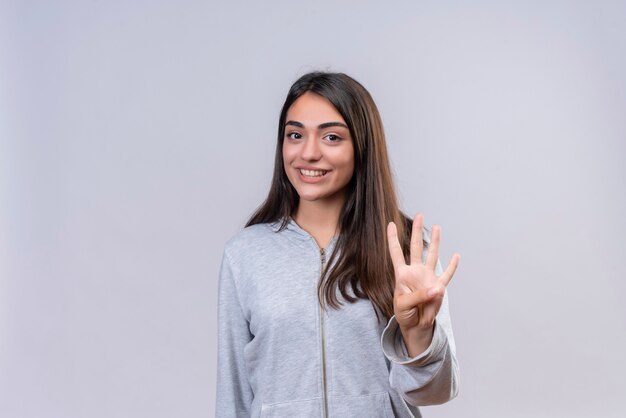 Hermosa joven mirando a la cámara con una sonrisa en la cara y mostrando cuatro números en la mano de pie sobre fondo blanco.