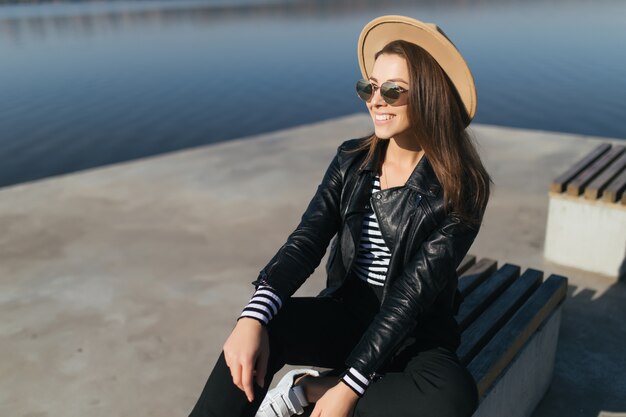 Hermosa joven modelo niña mujer sentada en un banco en día de otoño en la orilla del lago vestida con ropa casual