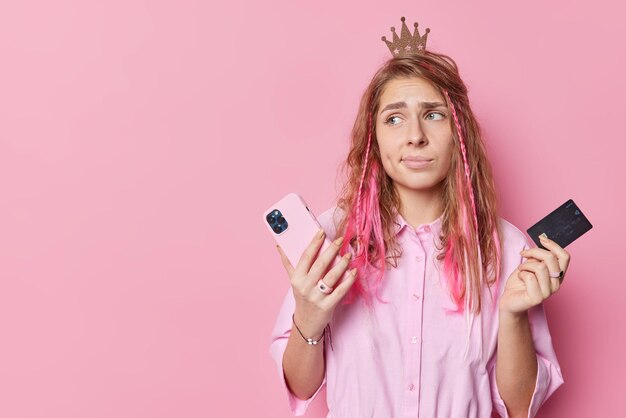 Una hermosa joven molesta se ve frustrada y no tiene dinero en pedidos de cuentas bancarias. Algo en línea tiene un teléfono inteligente y una tarjeta de crédito concentrados aislados sobre un espacio en blanco de fondo rosa.