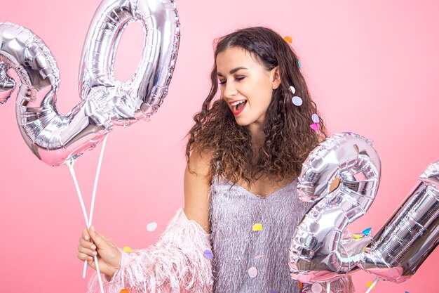 Hermosa joven morena con cabello rizado y ropa festiva posando sobre un fondo de estudio rosa con confeti y sosteniendo en su mano globos plateados para el concepto de año nuevo