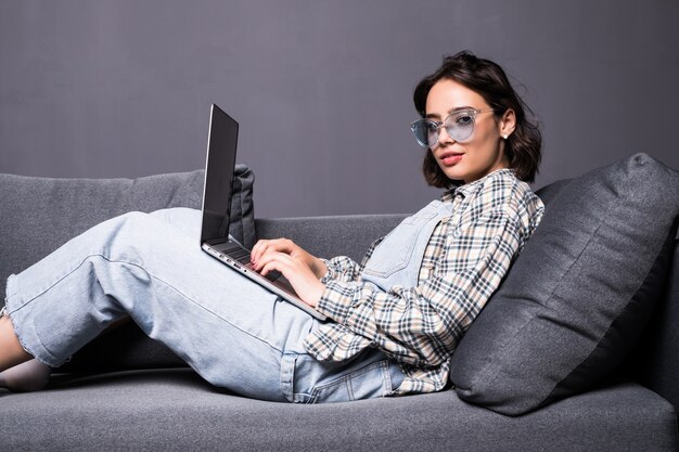 Hermosa joven morena en casa sentado en el sofá o sofá usando su computadora portátil y sonriendo