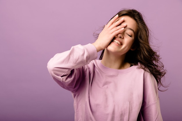 Foto gratuita hermosa joven morena caucásica sonriendo cubriendo la cara con la mano de pie sobre fondo púrpura concepto de buen humor