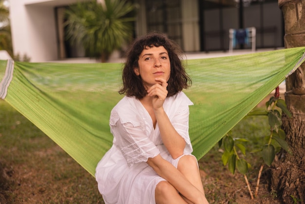 Hermosa joven morena caucásica con vestido blanco mira a la cámara sentada al aire libre en verano