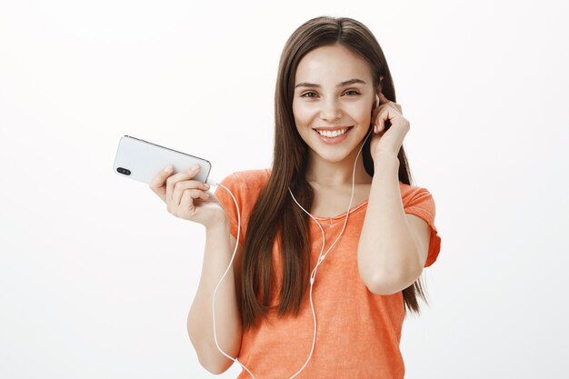 Hermosa joven morena escuchando música en auriculares y sosteniendo el teléfono móvil