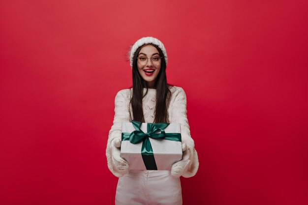 Hermosa joven morena con gafas, gorro de punto y guantes sonriendo y sosteniendo una caja de regalo de Navidad contra un fondo rojo liso