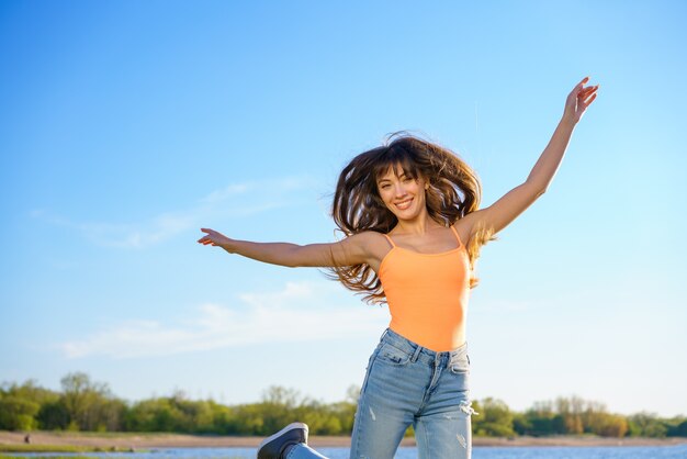 Una hermosa joven morena en jeans y una camiseta naranja salta contra el cielo en un día soleado de verano