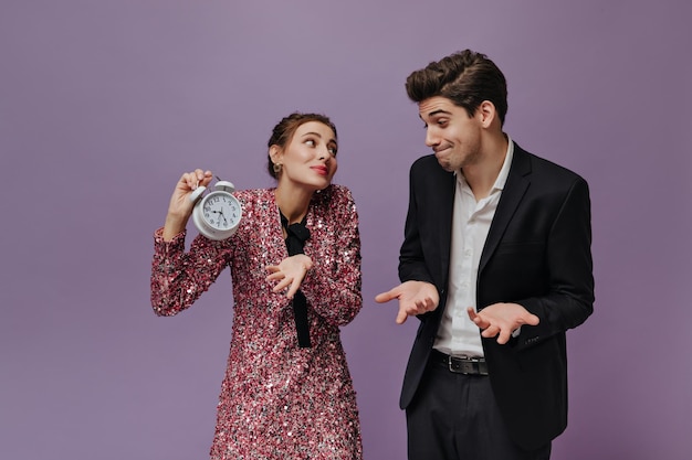 Hermosa joven morena con vestido rosa que muestra emocionalmente la hora en el reloj blanco a su novio con camisa y traje negro Par posando aislada en el fondo violeta