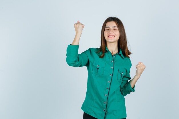 Hermosa joven mostrando gesto de ganador en camisa verde y mirando feliz, vista frontal.