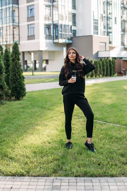 Hermosa joven mujer caucásica en traje deportivo negro y zapatillas negras va a caminar a la ciudad con una taza de café