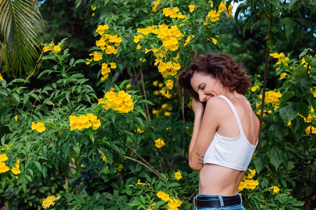 Hermosa joven mujer feliz caucásica ajuste elegante delgado en top blanco y pantalones de mezclilla en el parque rodeado de flores amarillas tailandesas