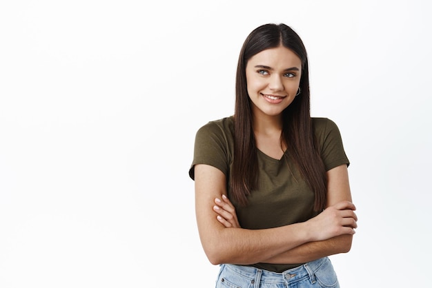 Hermosa joven mujer sana sonriendo mirando hacia otro lado en el espacio de la copia del logotipo de pie con los brazos cruzados sobre el pecho contra el fondo blanco con una camiseta informal