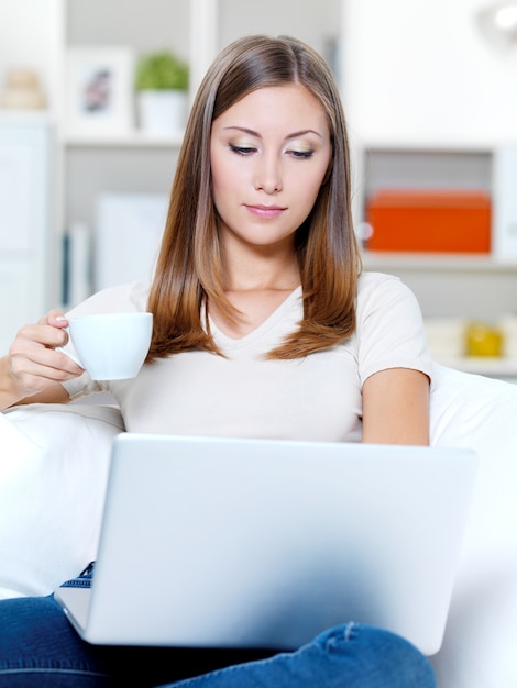 Hermosa joven mujer seria con laptop y taza de café en el sofá