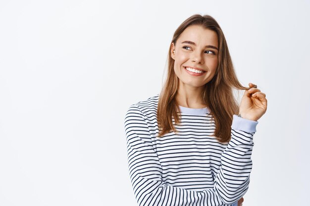 Hermosa joven mujer sonriente con dientes blancos, jugando con un mechón de cabello y mirando a la izquierda con rostro pensativo, mirando publicidad o logotipo en el espacio de la copia, blanco