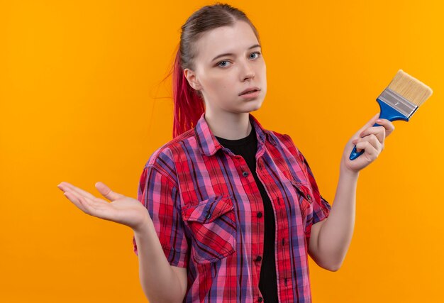 Hermosa joven mujer vistiendo camisa roja sosteniendo pincel sobre pared amarilla aislada