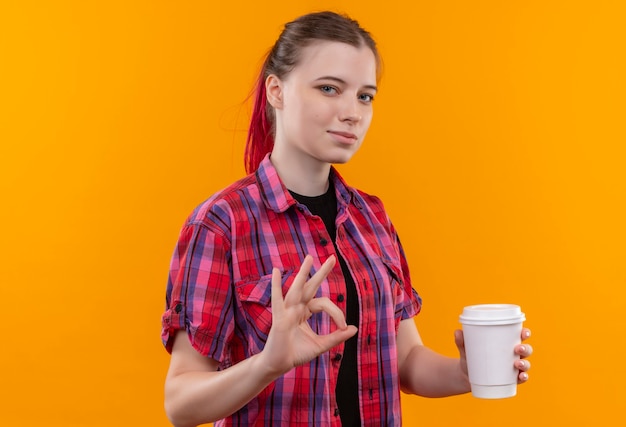 Hermosa joven mujer vistiendo camisa roja sosteniendo una taza de café mostrando gesto okey en pared amarilla aislada
