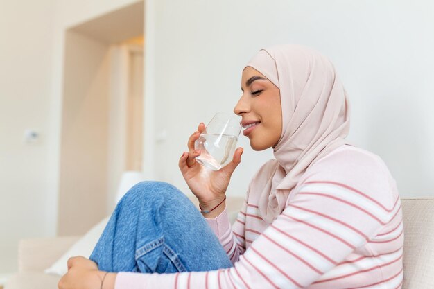 Hermosa joven musulmana con hiyab bebiendo un vaso de agua fresca en casa