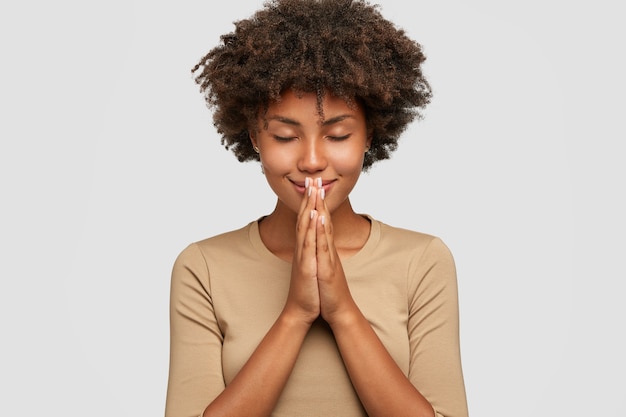 Hermosa joven negra se encuentra en pose meditativa, disfruta de un ambiente tranquilo, toma de la mano en gesto de oración