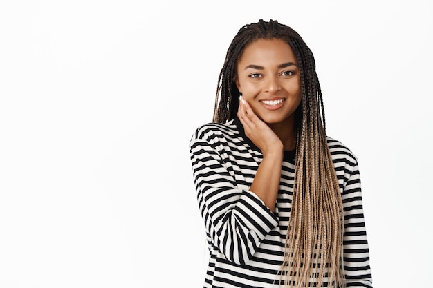 Hermosa joven negra con piel facial natural sin maquillaje tocándose la cara y sonriendo con placer de pie sobre fondo blanco