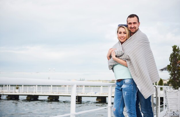 Hermosa joven pareja abrazándose, de pie en un muelle cerca del agua.