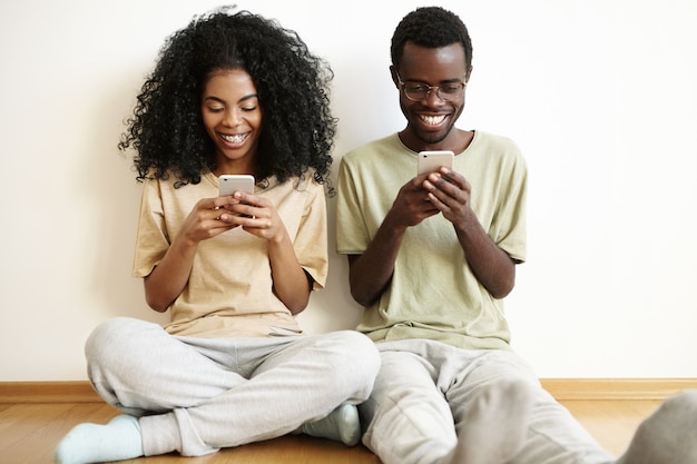 Hermosa joven pareja africana disfrutando de la comunicación en línea en casa, sentada en el piso, usando aparatos electrónicos