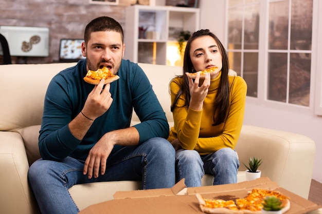 Hermosa joven pareja comiendo pizza mientras ve la televisión en la sala de estar sentado en el sofá