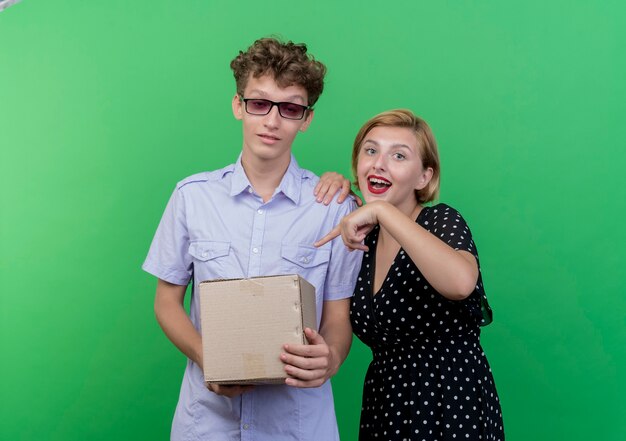 Hermosa joven pareja confundido hombre sosteniendo el paquete de caja mientras su novia apuntando con el dedo a la caja feliz sonriendo de pie sobre la pared verde