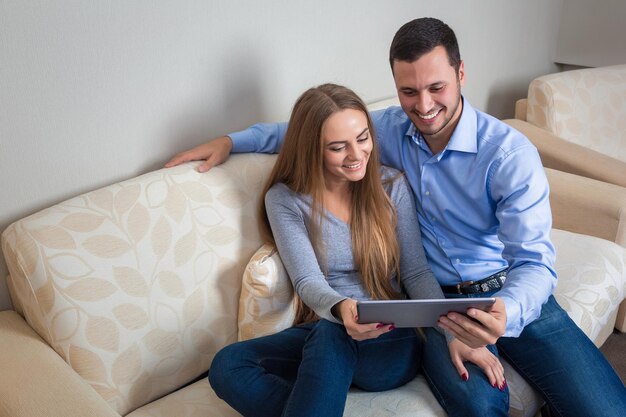 Hermosa joven pareja encantadora, riéndose sentada en un sofá, compartiendo fotos u otra información, mostrada en una tableta electrónica entre ellos