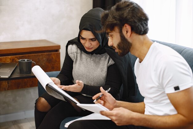 Hermosa joven pareja escribiendo en un cuaderno, sentado en la camilla en casa. Chica árabe con hidjab negro.