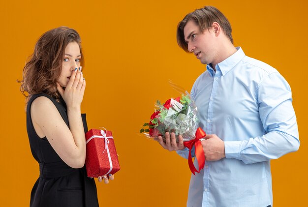 Hermosa joven pareja hombre feliz con ramo de rosas rojas mirando a su novia sorprendida con presente celebrando el día de San Valentín de pie sobre fondo naranja
